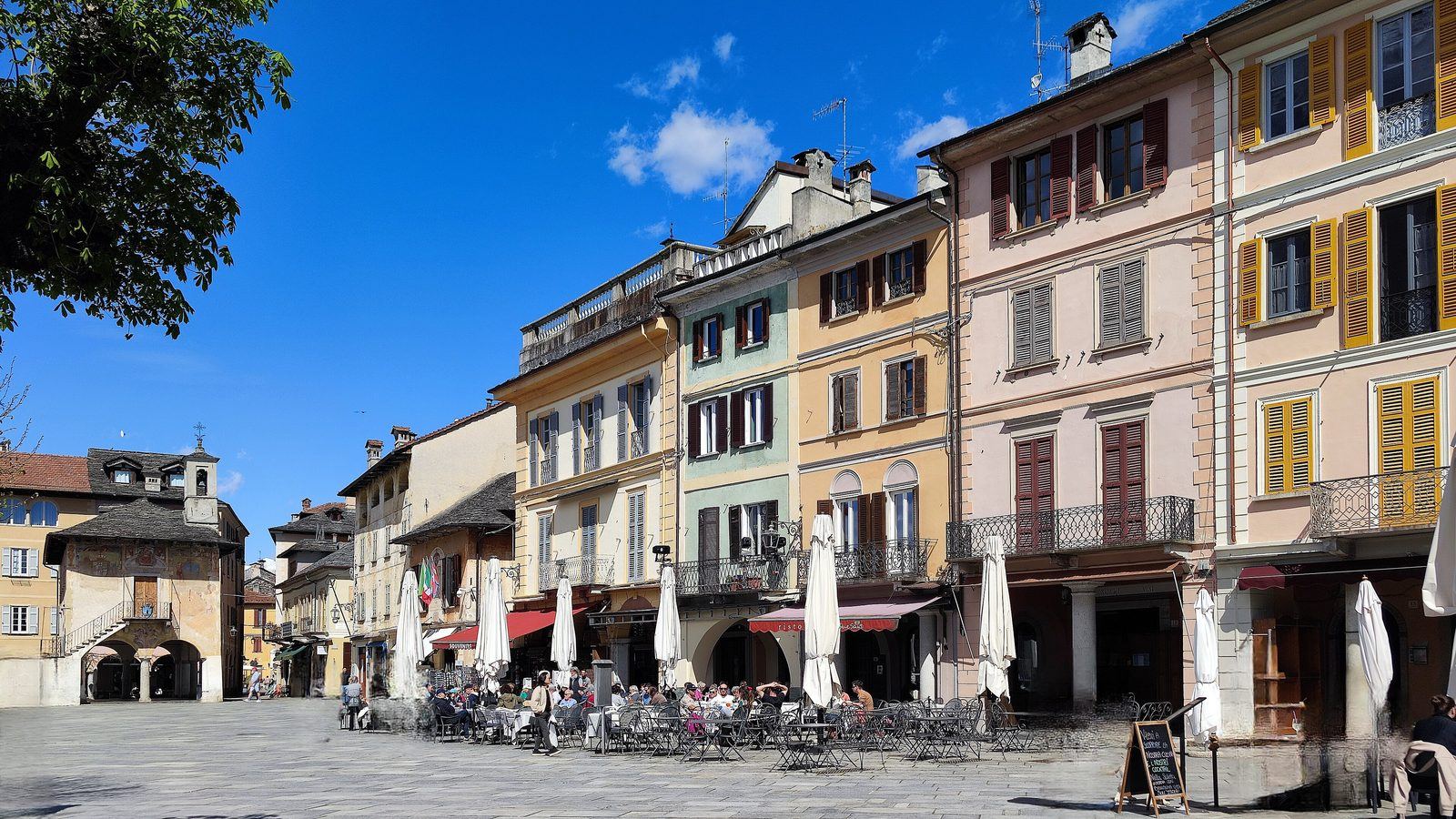 Café-Tische und Sonnenschirme auf der Piazza Motta in Orta San Giulio vor pastellfarbenen Palazzi mit Arkaden