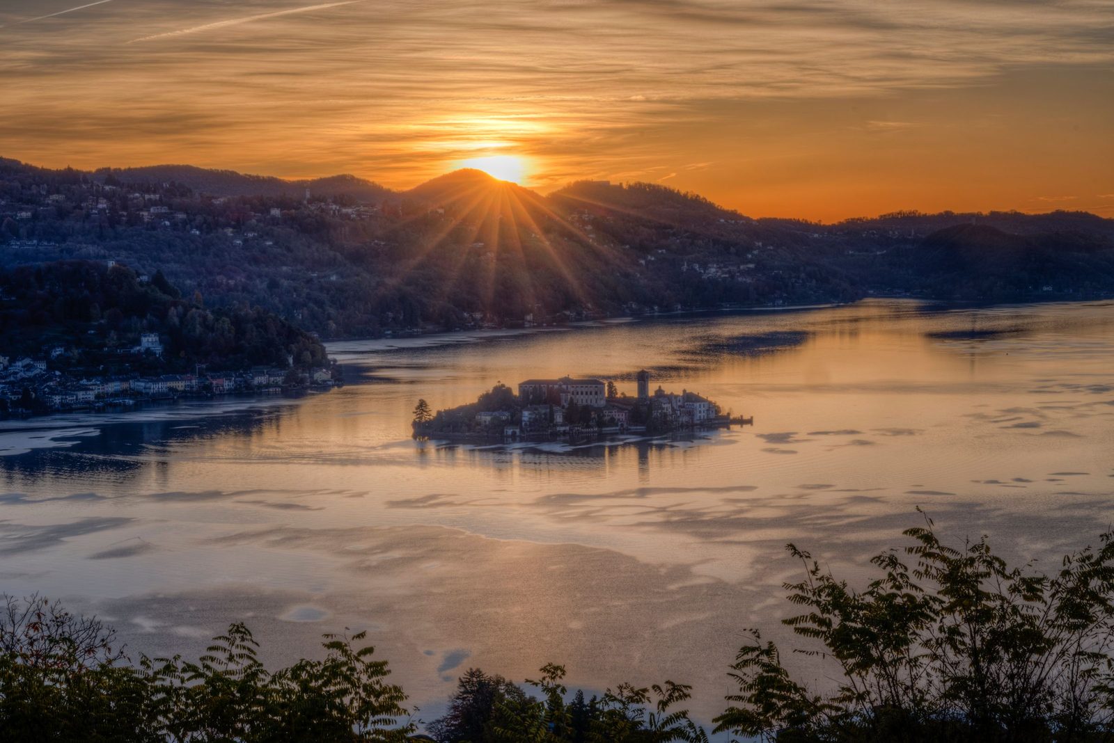 Die Sonne geht hinter den Hügeln über dem Ortasee unter, die Isola San Giulio als Silhouette auf goldenem Wasser